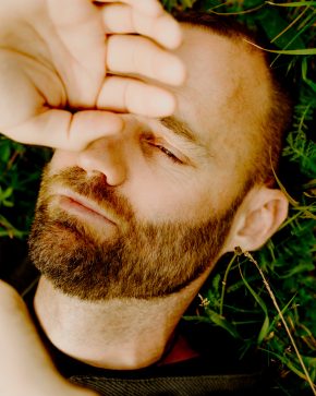 Close-up portrait of Thor Pedersen lying in the grass, shielding his eyes from the sun with his hand.