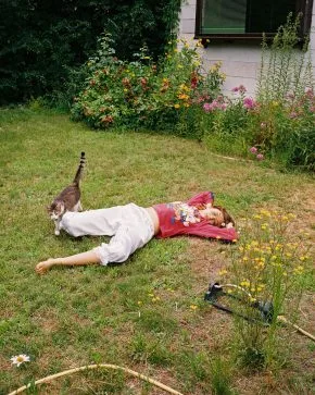 woman laying on the grass in a backyard while a cat walks by